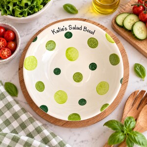 May include: A round, cream-coloured ceramic bowl featuring a hand-painted pattern of various shades of green polka dots. The text "Katie's Salad Bowl" is printed in black lettering along the upper inner rim of the bowl, which sits on a wooden board.