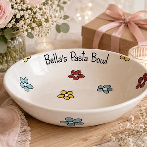 May include: A white ceramic pasta bowl featuring hand-painted colourful flowers in red, yellow, and blue. The interior rim is inscribed with the black text Bella's Pasta Bowl. The bowl has a smooth, glossy finish and a wide, shallow circular shape.