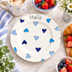 May include: A round white ceramic plate featuring a hand-painted pattern of scattered light and dark blue hearts. The name "Halle" is written in black script at the top of the plate. The design is simple, clean, and features a smooth, glossy finish.