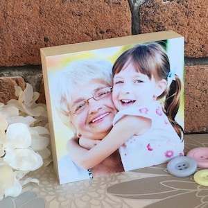 May include: A wooden photo block with a picture of an older woman and a young girl smiling and hugging. The girl is wearing a white and pink floral dress.