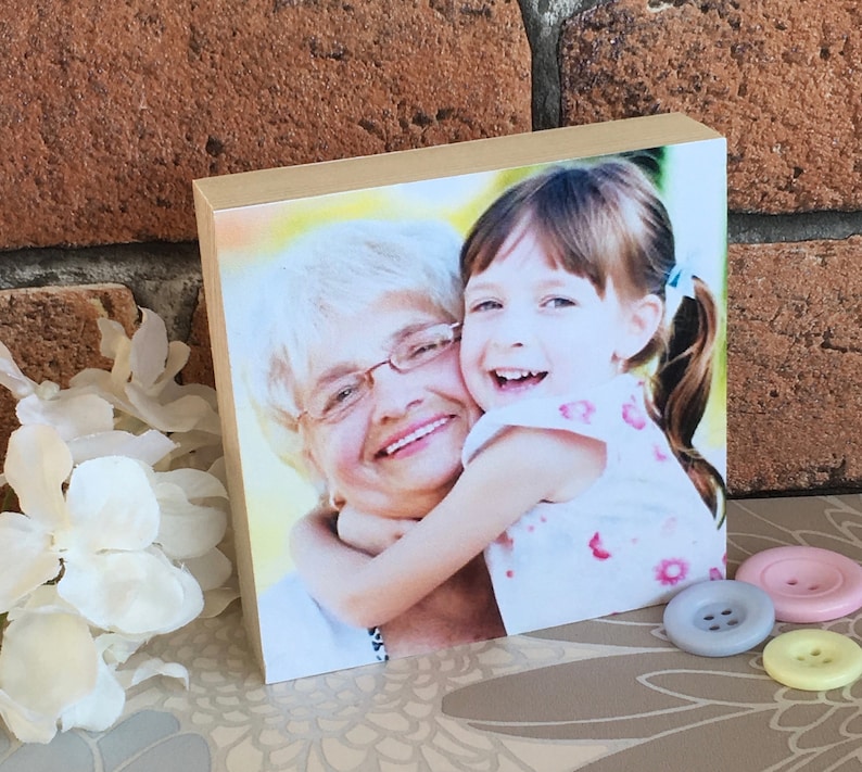 May include: A wooden photo block with a picture of an older woman and a young girl smiling and hugging. The girl is wearing a white and pink floral dress.