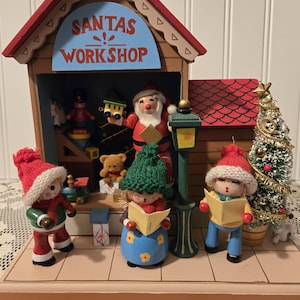 May include: A Christmas-themed wooden display featuring a Santa's Workshop scene. The display includes figurines of carolers, Santa Claus, and a Christmas tree. The building has a red roof and the words "SANTAS WORKSHOP" in blue.