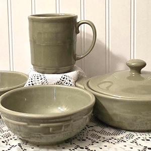 May include: A collection of olive green stoneware kitchenware, including a mug, two bowls, and a lidded casserole dish. The mug is cylindrical with a handle. The bowls have a textured design. The casserole dish has a round lid with a knob.