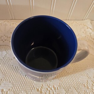 May include: A white ceramic mug with a blue interior and a white handle. The mug has a simple design with a blue rim and a blue interior. The mug is sitting on a white lace tablecloth.