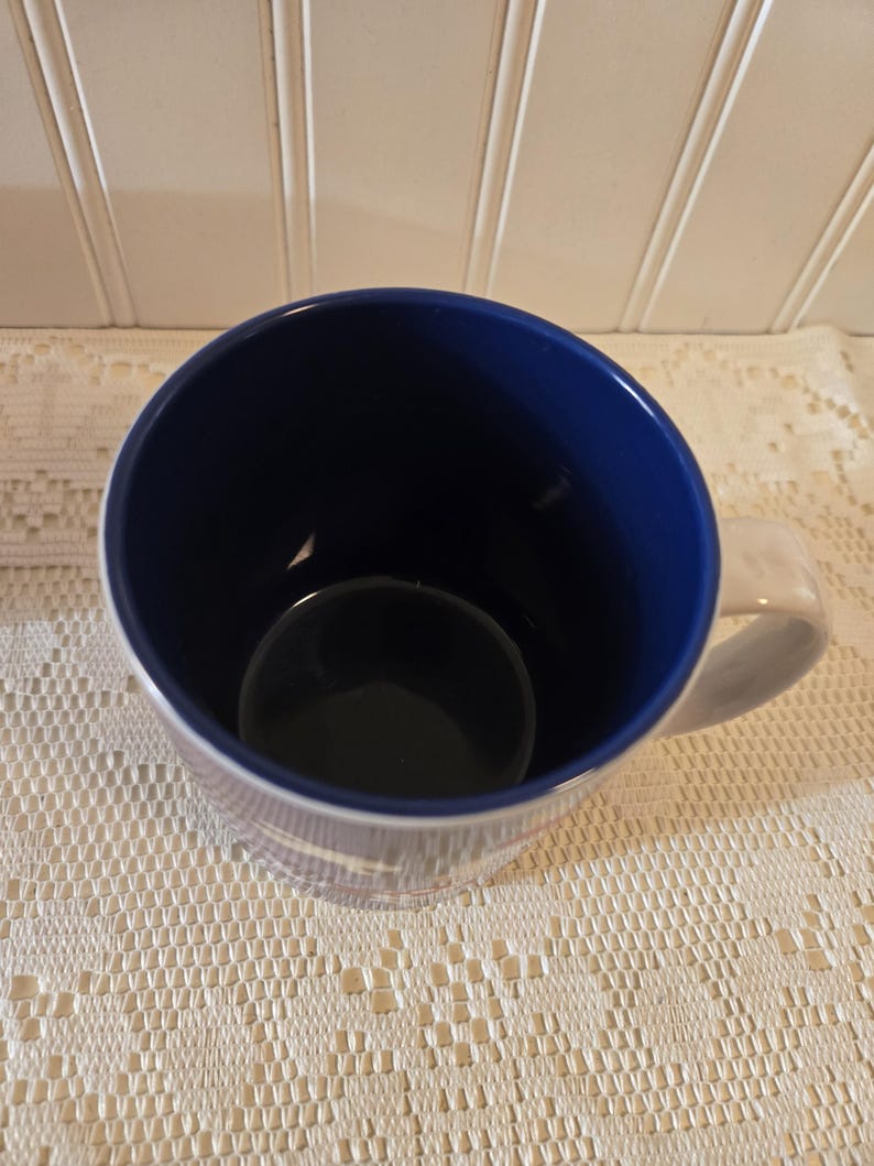 May include: A white ceramic mug with a blue interior and a white handle. The mug has a simple design with a blue rim and a blue interior. The mug is sitting on a white lace tablecloth.