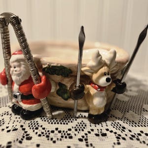 May include: A ceramic Christmas-themed nut bowl with Santa Claus and a reindeer on either side. Santa holds a nutcracker, and the reindeer holds two nut picks. The bowl is beige with green leaf accents.