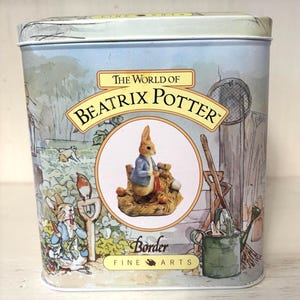 May include: A rectangular tin featuring illustrations from "The World of Beatrix Potter." The tin has a light blue background with images of rabbits, a bird, and garden tools. The center of the tin features a circular image of a rabbit wearing a blue jacket.