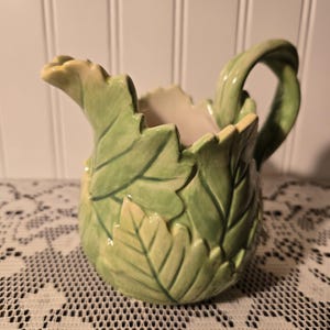 May include: A ceramic creamer pitcher with a leaf design. The pitcher is primarily green with lighter green and yellow accents. The handle is a twisted vine shape.