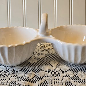 May include: White ceramic serving dish with two connected bowls and a handle. The bowls have a scalloped rim and vertical ribbed sides. The dish is on a white lace tablecloth, with a white paneled wall in the background.