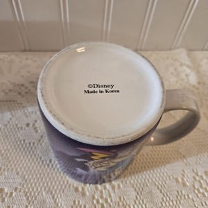 May include: A white ceramic mug with a gray handle, featuring a purple and blue design. The bottom of the mug has the text "&copy;Disney Made in Korea". The mug is sitting on a white lace tablecloth.