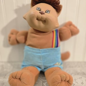 May include: A plush cat doll with tan fabric skin, brown yarn hair, and embroidered facial features. The doll wears light blue shorts with a rainbow-striped suspender strap over the left shoulder and has visible paw-like feet and hands.