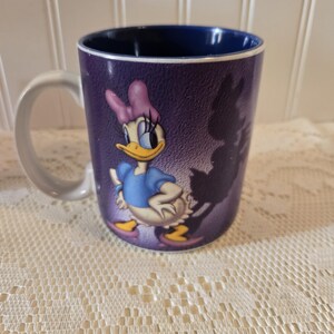 May include: A ceramic mug featuring Daisy Duck from Disney. The mug has a purple exterior with a graphic of Daisy Duck in a blue dress and pink bow. The interior of the mug is dark blue, and the handle is white.