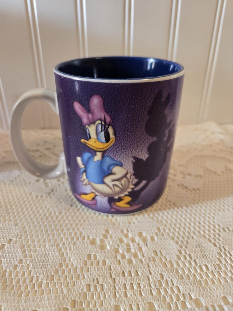May include: A ceramic mug featuring Daisy Duck from Disney. The mug has a purple exterior with a graphic of Daisy Duck in a blue dress and pink bow. The interior of the mug is dark blue, and the handle is white.