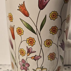 May include: White ceramic mug decorated with colorful floral illustrations. The design features orange, purple, yellow, and pink flowers with green leaves and black stems. The mug has a white handle.
