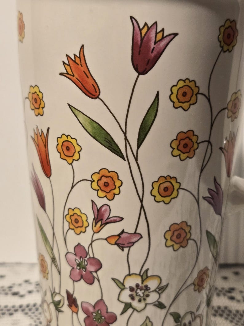 May include: White ceramic mug decorated with colorful floral illustrations. The design features orange, purple, yellow, and pink flowers with green leaves and black stems. The mug has a white handle.