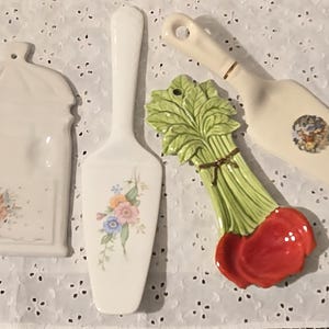 May include: Four ceramic kitchen utensils: a white utensil holder, a cake server, a radish-shaped trivet, and a cake server. The utensils are decorated with floral patterns and are set against a white, patterned background.