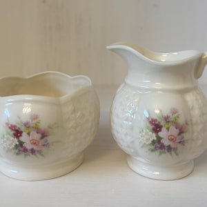May include: Cream-colored ceramic creamer and sugar bowl set. The creamer has a rounded body and a handle. The sugar bowl is round with a scalloped rim. Both pieces feature a floral design with pink, purple, and yellow flowers.