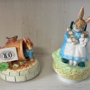 May include: Two ceramic figurines of Peter Rabbit characters. One features Peter Rabbit emerging from a flower pot with a sign that reads "Peter Rabbit 100 Schmid 1993". The other shows a rabbit in a blue dress holding two smaller rabbits.