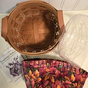 May include: A Longaberger basket set with a round, woven brown basket, leather handles, a clear plastic liner, and a floral fabric liner. The fabric liner features a pattern of red, yellow, and orange leaves.