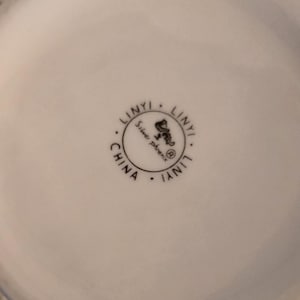 K&ouml;nnte beinhalten: Nahaufnahme eines wei&szlig;en Keramikobjekts mit einem runden Stempel. Der Stempel lautet "LINYI" am Umfang, mit "Silver phoenix" und einer Phoenix-Illustration in der Mitte. Das Wort "CHINA" ist ebenfalls vorhanden.