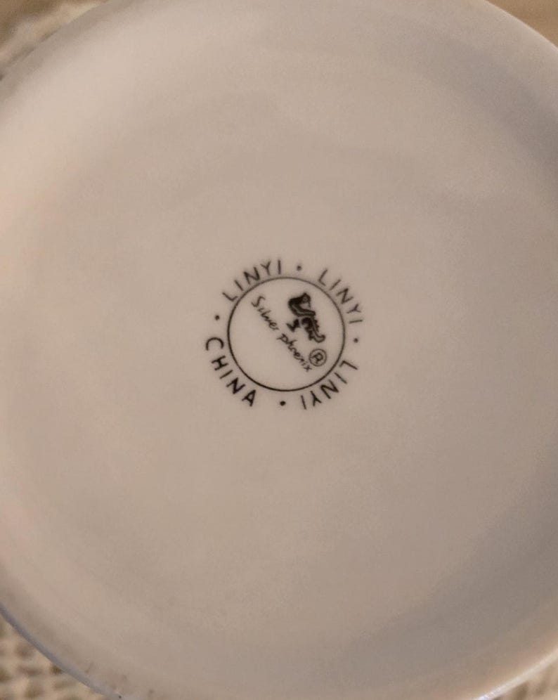 K&ouml;nnte beinhalten: Nahaufnahme eines wei&szlig;en Keramikobjekts mit einem runden Stempel. Der Stempel lautet "LINYI" am Umfang, mit "Silver phoenix" und einer Phoenix-Illustration in der Mitte. Das Wort "CHINA" ist ebenfalls vorhanden.