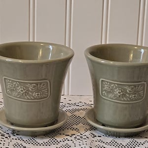 May include: Two sage green ceramic flower pots with matching saucers. Each pot has a rectangular embossed floral design. The pots are on a white lace tablecloth against a white paneled wall.