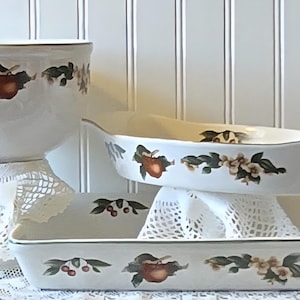 May include: A set of three white ceramic kitchen vessels including a deep round bowl, an oval baking dish with side handles, and a rectangular baking pan. Each piece features a repeating pattern of red apples, green leaves, and white blossoms.