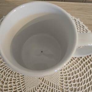 K&ouml;nnte beinhalten: Eine wei&szlig;e Keramiktasse mit einem abgerundeten Henkel. Die Tasse ist leer und steht auf einer wei&szlig;en, offen gewebten Tischdecke. Die Tasse wird von oben betrachtet und zeigt das Innere und den Henkel.
