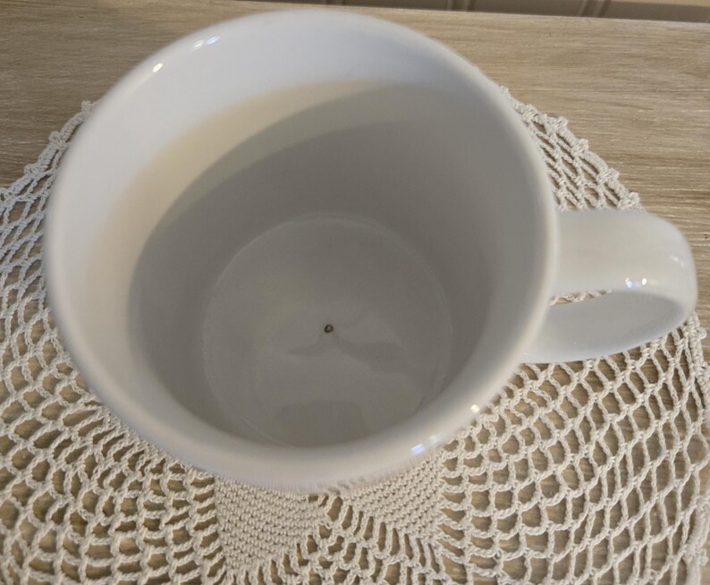 K&ouml;nnte beinhalten: Eine wei&szlig;e Keramiktasse mit einem abgerundeten Henkel. Die Tasse ist leer und steht auf einer wei&szlig;en, offen gewebten Tischdecke. Die Tasse wird von oben betrachtet und zeigt das Innere und den Henkel.