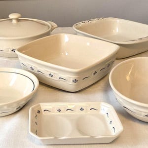 May include: A collection of cream-colored ceramic kitchenware featuring a dark blue decorative border pattern. The set includes a lidded casserole dish, a rectangular baking pan, a square serving bowl, two round bowls, and a small rectangular tray.