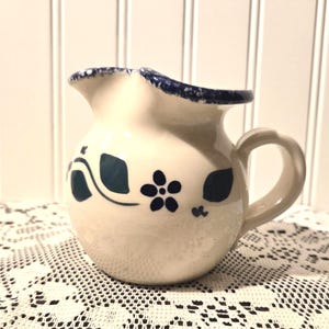 May include: A small, off-white ceramic pitcher with a rounded body and a curved handle. The pitcher features dark blue floral and leaf designs, with a dark blue trim around the spout.