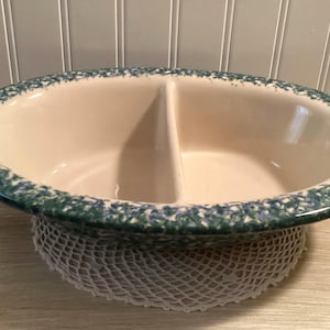 May include: Oval, two-section serving dish with a cream-colored interior and a speckled blue and green rim. The dish is made of ceramic and sits on a white, crocheted doily. A divider separates the two sections.
