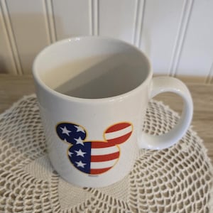K&ouml;nnte beinhalten: Wei&szlig;e Keramiktasse mit einem Mickey Mouse-Kopf-Design im patriotischen Stil. Das Design zeigt die amerikanische Flagge mit roten und wei&szlig;en Streifen und blauen Sternen. Die Tasse hat einen gebogenen Griff und steht auf einer wei&szlig;en Spitzendecke.