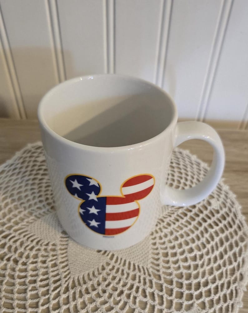 K&ouml;nnte beinhalten: Wei&szlig;e Keramiktasse mit einem Mickey Mouse-Kopf-Design im patriotischen Stil. Das Design zeigt die amerikanische Flagge mit roten und wei&szlig;en Streifen und blauen Sternen. Die Tasse hat einen gebogenen Griff und steht auf einer wei&szlig;en Spitzendecke.