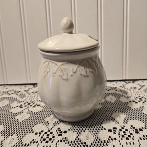 May include: A cream-colored ceramic jar with a lid and a small knob on top. The jar has a rounded shape with a leaf design around the middle. The jar is sitting on a white lace tablecloth.
