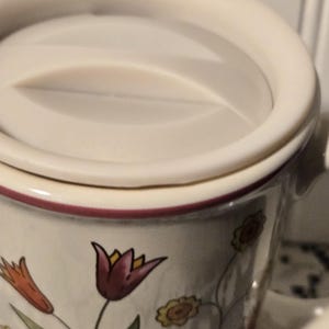 May include: Cream-colored ceramic mug with a matching lid. The mug is decorated with floral designs in shades of orange, yellow, and burgundy. The rim of the mug and lid are trimmed with a burgundy band.