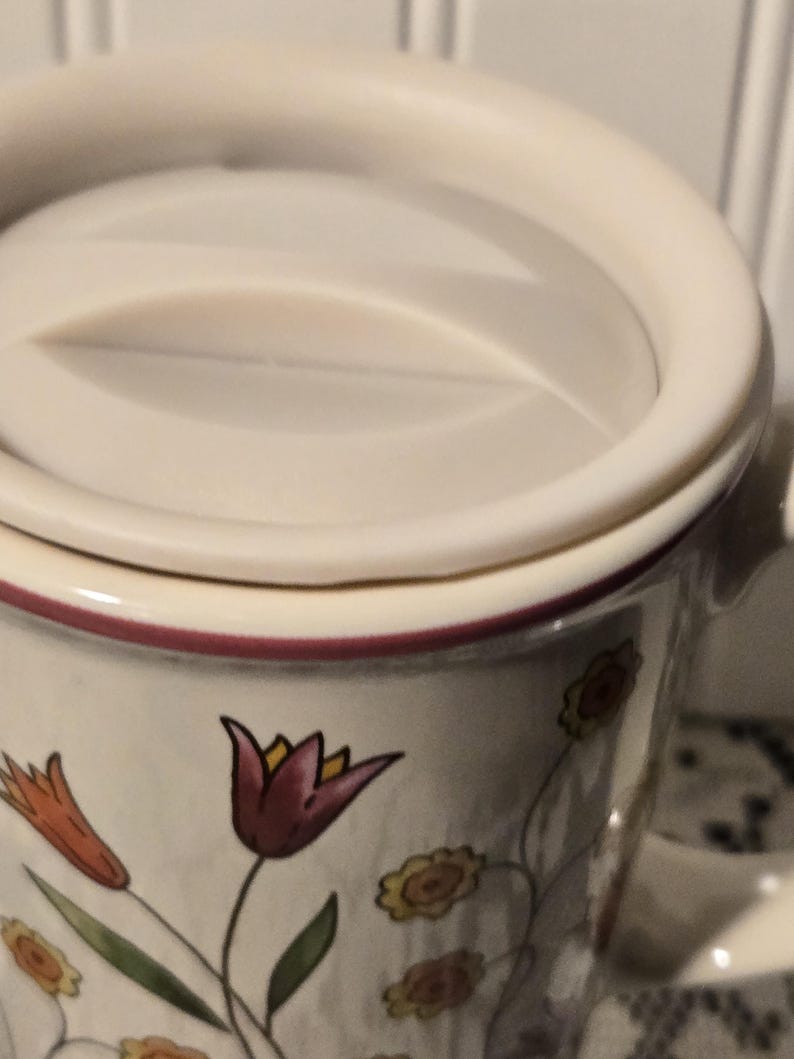 May include: Cream-colored ceramic mug with a matching lid. The mug is decorated with floral designs in shades of orange, yellow, and burgundy. The rim of the mug and lid are trimmed with a burgundy band.
