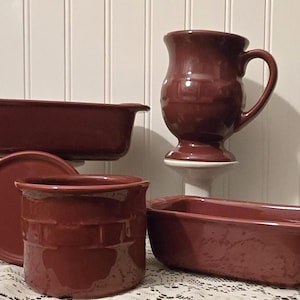 May include: A collection of maroon ceramic kitchenware, including a rectangular baking dish, a round plate, a cylindrical container, a small loaf pan, and a handled mug. The pieces are arranged on a white surface.