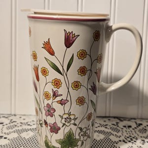 May include: A tall, off-white ceramic travel mug with a handle and a lid. The mug is decorated with a floral pattern featuring orange, yellow, and purple flowers with green leaves. The rim is trimmed with a thin red line.