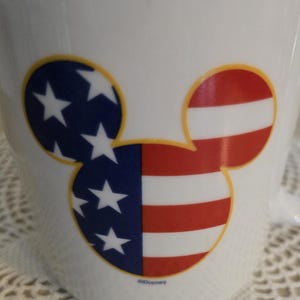K&ouml;nnte beinhalten: Wei&szlig;e Keramiktasse mit einem Mickey Mouse-Kopf-Design. Ein Ohr zeigt Blau mit wei&szlig;en Sternen, das andere hat rote und wei&szlig;e Streifen, die der amerikanischen Flagge &auml;hneln. Das Design ist gelb umrandet.