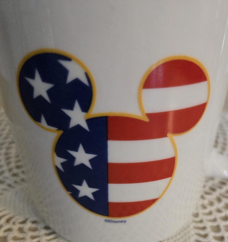 K&ouml;nnte beinhalten: Wei&szlig;e Keramiktasse mit einem Mickey Mouse-Kopf-Design. Ein Ohr zeigt Blau mit wei&szlig;en Sternen, das andere hat rote und wei&szlig;e Streifen, die der amerikanischen Flagge &auml;hneln. Das Design ist gelb umrandet.