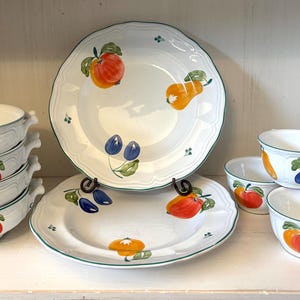 May include: White ceramic dishware set with fruit designs. Includes bowls, plates, and cups decorated with apples, pears, and plums. The set has a green trim and is arranged on a white shelf.