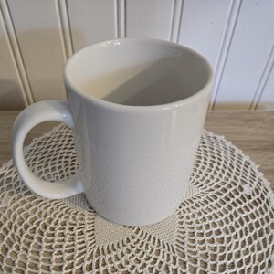 K&ouml;nnte beinhalten: Eine wei&szlig;e Keramik-Kaffeetasse mit gebogenem Henkel. Die Tasse steht auf einer cremefarbenen, geh&auml;kelten Untersetzer. Der Hintergrund ist eine helle Holzoberfl&auml;che und wei&szlig;e vertikale Paneele.