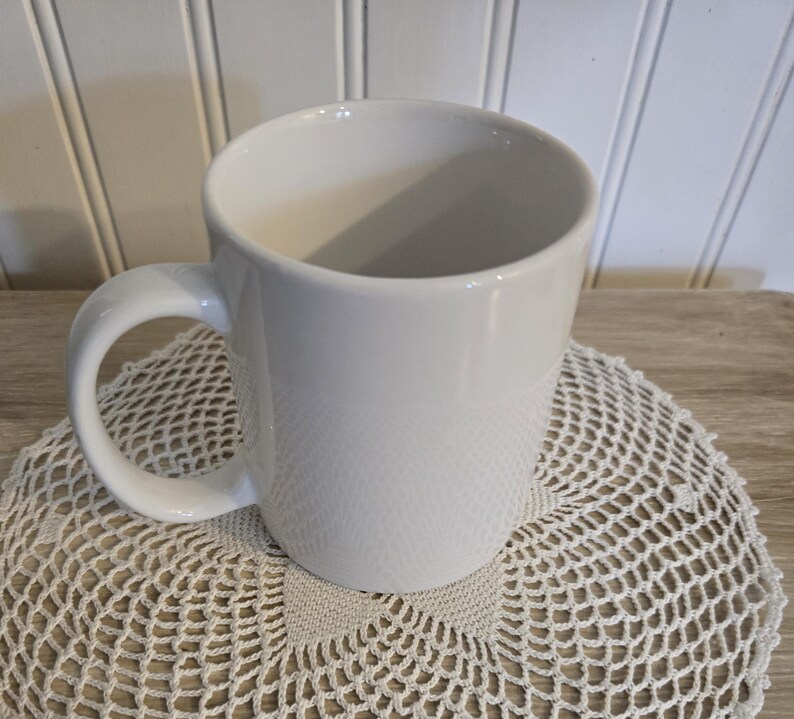 K&ouml;nnte beinhalten: Eine wei&szlig;e Keramik-Kaffeetasse mit gebogenem Henkel. Die Tasse steht auf einer cremefarbenen, geh&auml;kelten Untersetzer. Der Hintergrund ist eine helle Holzoberfl&auml;che und wei&szlig;e vertikale Paneele.
