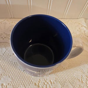 May include: A white ceramic mug with a blue interior and a white handle. The mug has a simple design and is sitting on a white lace tablecloth. The mug is empty.