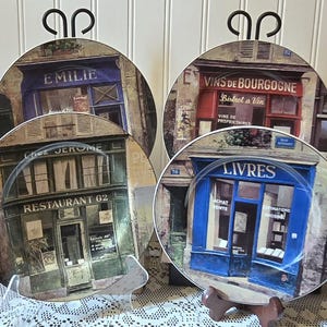 May include: Four decorative plates with images of French storefronts. The plates feature the names "EMILIE", "VINS DE BOURGOGNE", "RESTAURANT 62", and "LIVRES". Each plate is displayed on a decorative stand, showcasing the detailed artwork.