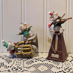 May include: Two Christmas ornaments featuring mice dressed in festive attire. One mouse is playing a violin atop a brown metronome base, while the other two are playing a brass instrument. The ornaments are from 1995.