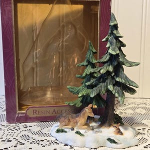 May include: A resin Christmas scene featuring two evergreen trees, a deer, and a fawn resting on a snowy base. The scene is part of the "Santa's Workbench Collection" and comes in a decorative box.