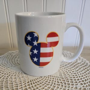K&ouml;nnte beinhalten: Wei&szlig;e Keramiktasse mit einem Mickey-Mouse-Kopf-Design im patriotischen Stil. Das Design zeigt die amerikanische Flagge mit roten und wei&szlig;en Streifen und blauen Sternen. Die Tasse hat einen gebogenen Griff.
