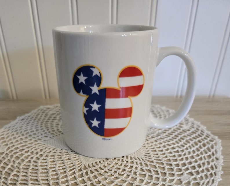 K&ouml;nnte beinhalten: Wei&szlig;e Keramiktasse mit einem Mickey-Mouse-Kopf-Design im patriotischen Stil. Das Design zeigt die amerikanische Flagge mit roten und wei&szlig;en Streifen und blauen Sternen. Die Tasse hat einen gebogenen Griff.